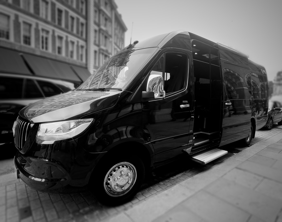 A black Mercedes-Benz Sprinter luxury minibus with its side door and step open on a Central London street, in monochrome.